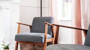 A chair with wooden armrests and a grey upholstered seat in a room with a wooden floor, a potted plant, and a window with curtains. - Stylish 4BR Family Home | Heart of Grimsby (Grimsby)