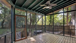 A covered outdoor deck with a wooden floor, a ceiling fan, and large windows offering views of a backyard with a hammock and patio furniture.