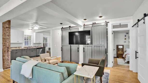 A spacious living room with a sofa, a coffee table, and a TV. There is a kitchen area with cabinets and a dining table.
