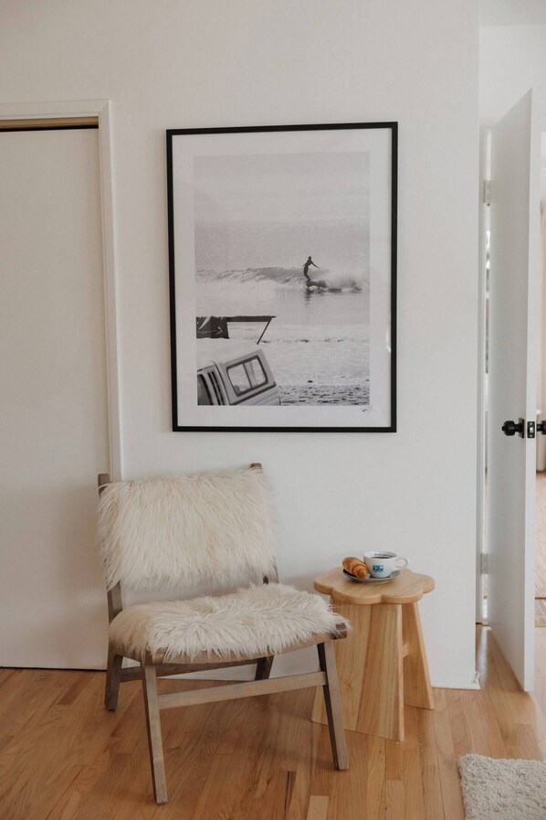 A room with a wooden chair, a small wooden stool with a cup and a croissant, and a framed black and white photograph on the wall.