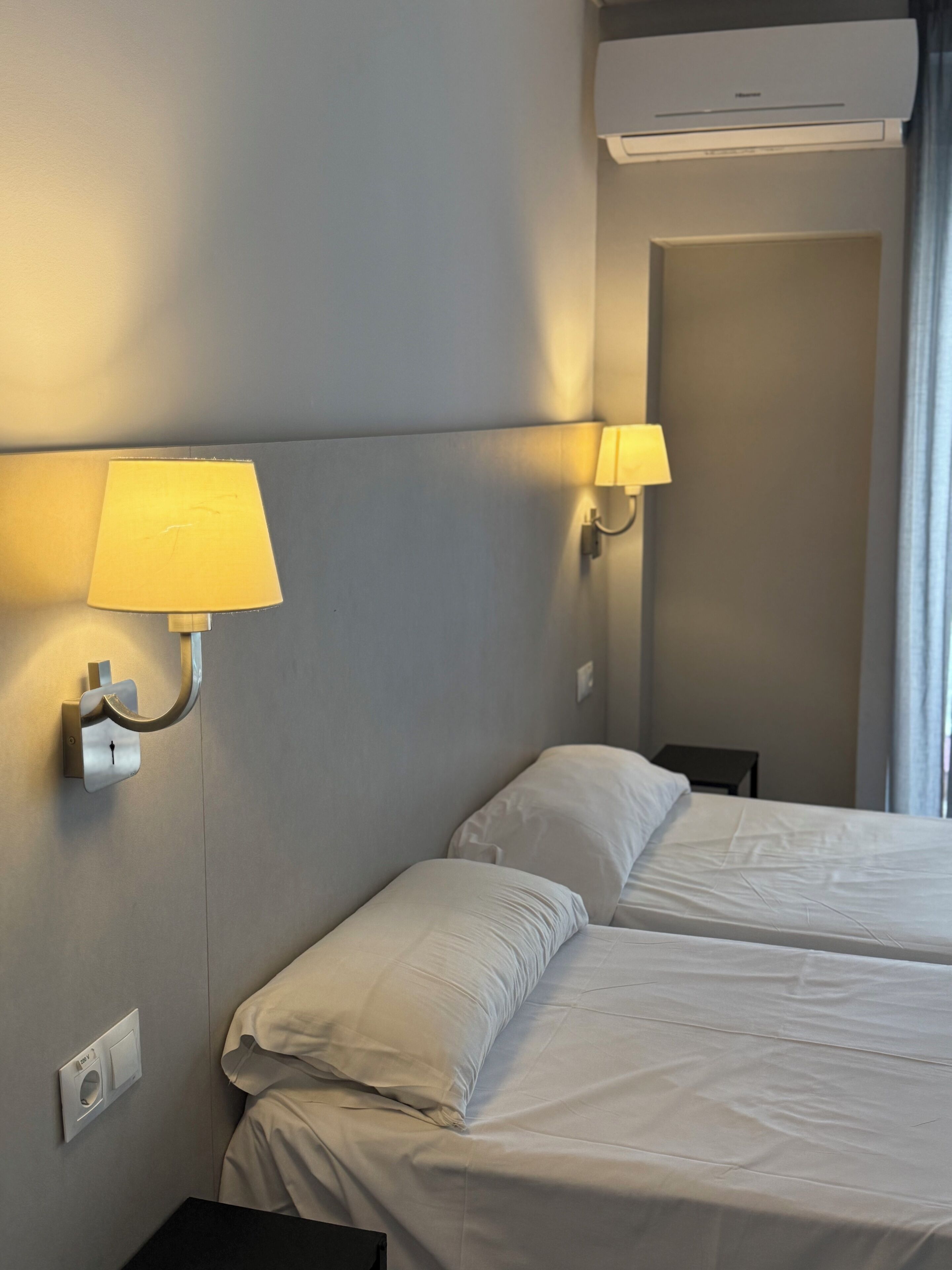 Ein Hotelzimmer mit zwei Betten, einer Wandleuchte, einer Klimaanlage und einer Steckdose.