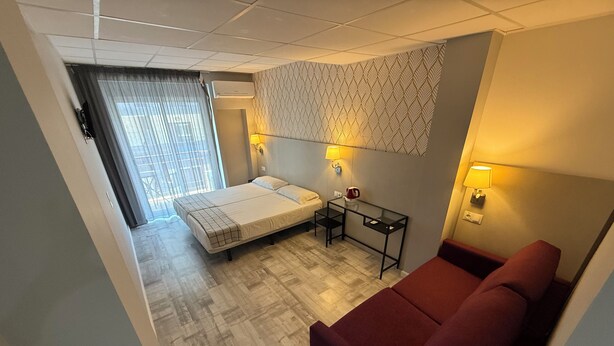 A hotel room with a bed, a small table, a red sofa, a patterned wall, and a window with curtains. - Hotel B and B Suites (Benidorm)