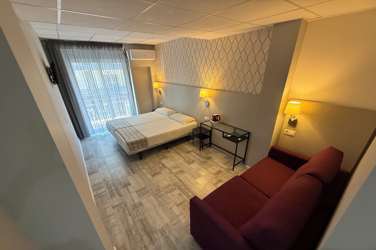 A hotel room with a bed, a small table, a red sofa, a patterned wall, and a window with curtains.