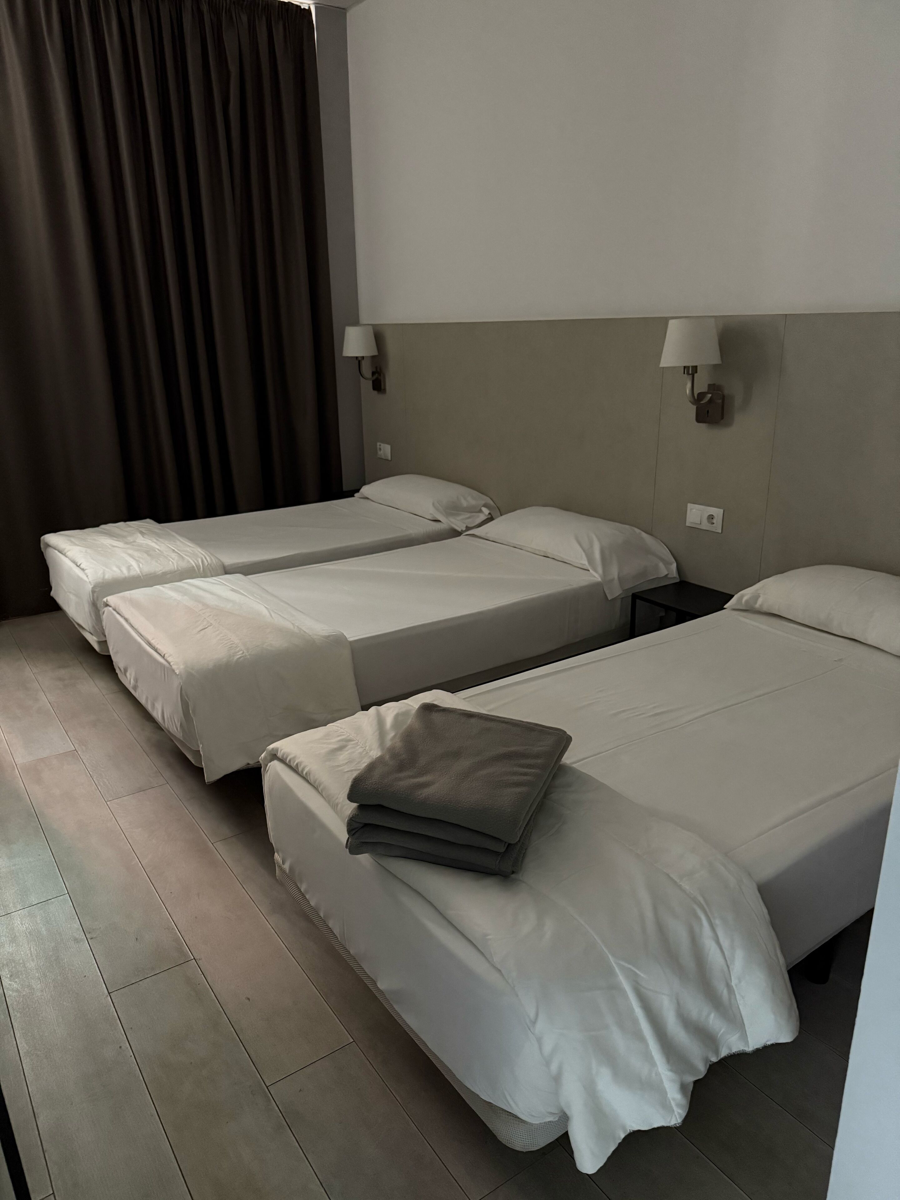 Una habitación de hotel con dos camas individuales, una cortina oscura, una lámpara de pared y una mesita.