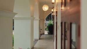 Un couloir avec un ventilateur de plafond, des luminaires suspendus et une vue sur un jardin où poussent des fleurs roses.
