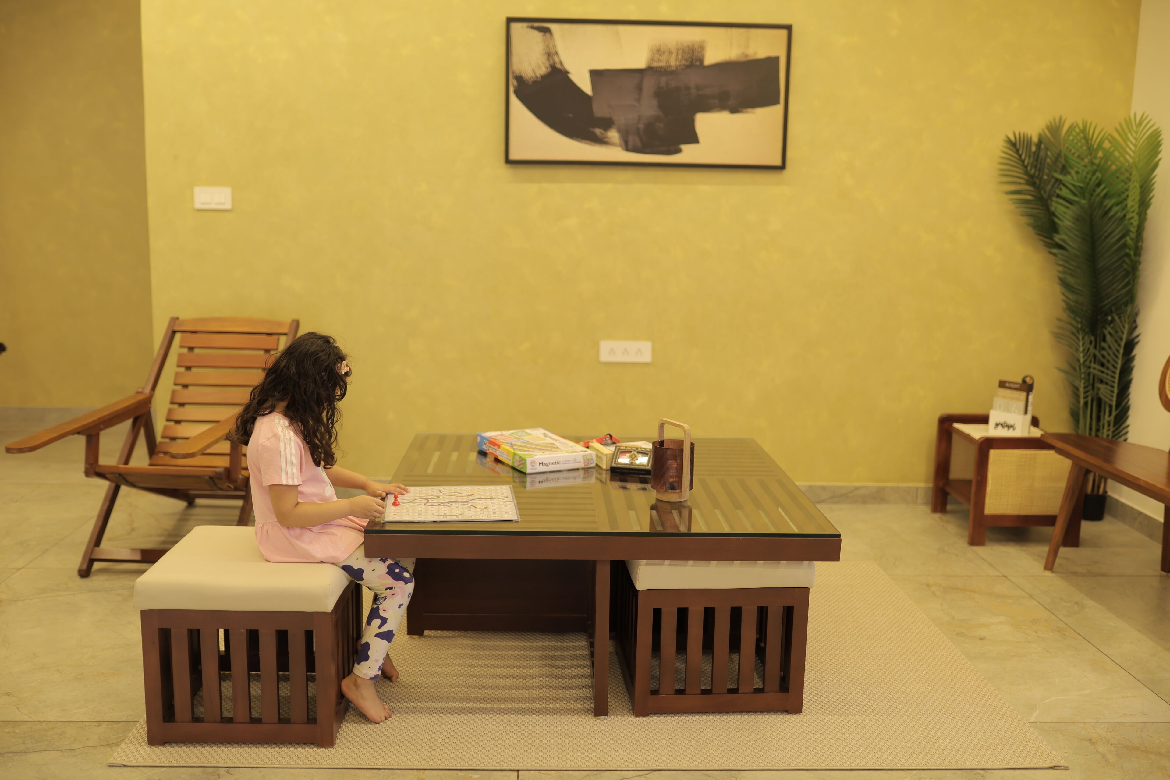 A child sitting at a table with a book, surrounded by wooden furniture and a large plant.