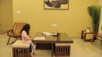 A child sitting at a table with a book, surrounded by wooden furniture and a large plant.