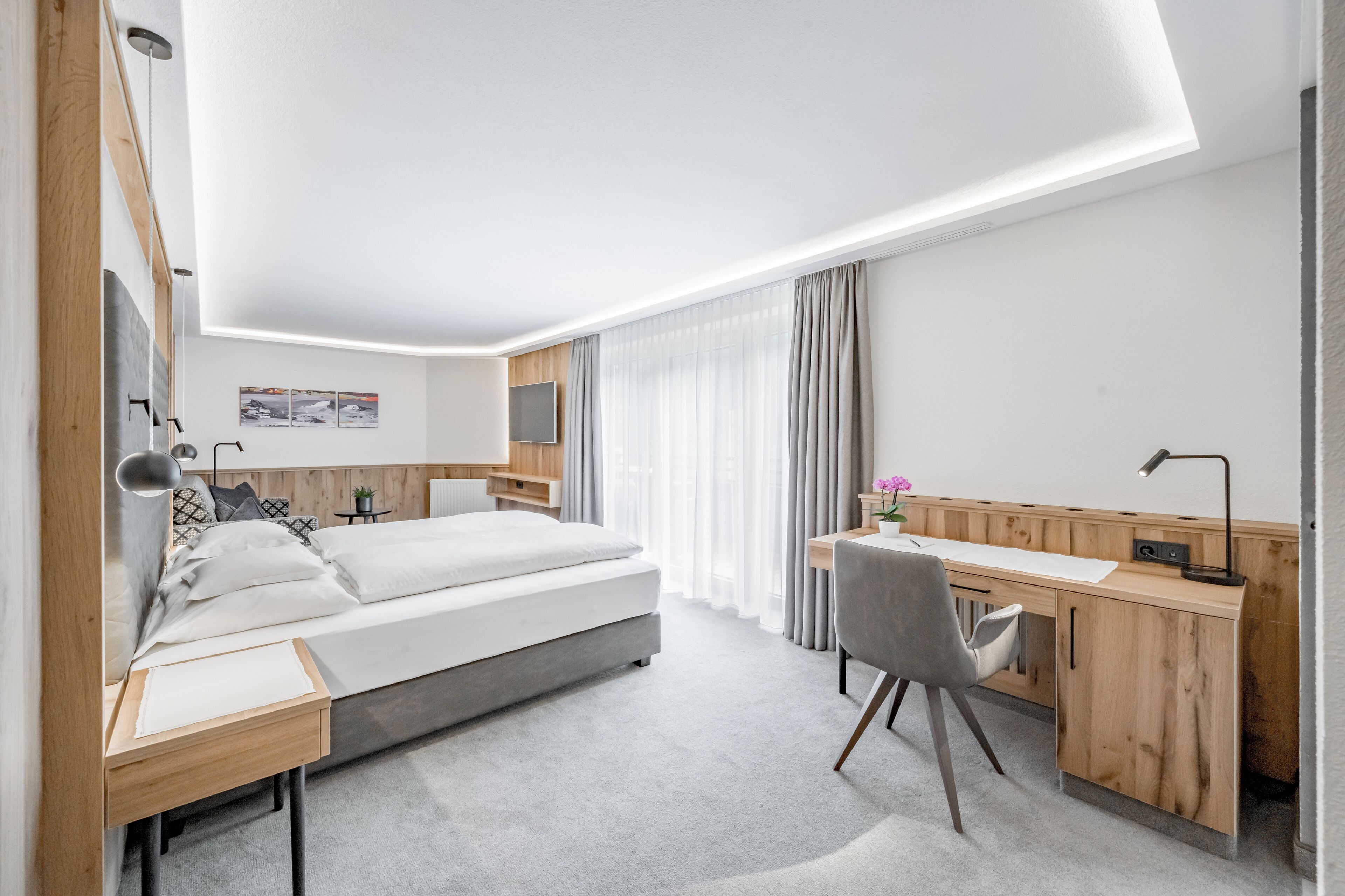 A modern hotel room with a bed, desk, and chair, featuring a wooden desk and a grey chair.