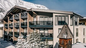 Un bâtiment moderne à plusieurs étages, doté de balcons, avec un arbre recouvert de neige et une petite structure ressemblant à une tour.