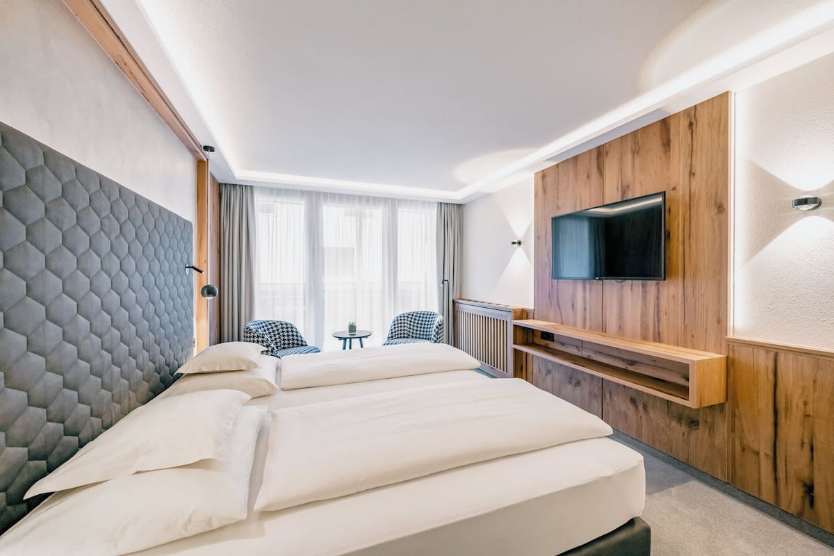 Habitación de hotel con una cama grande, un televisor instalado en una pared de paneles de madera, una pequeña mesa con sillas y una ventana con cortinas.