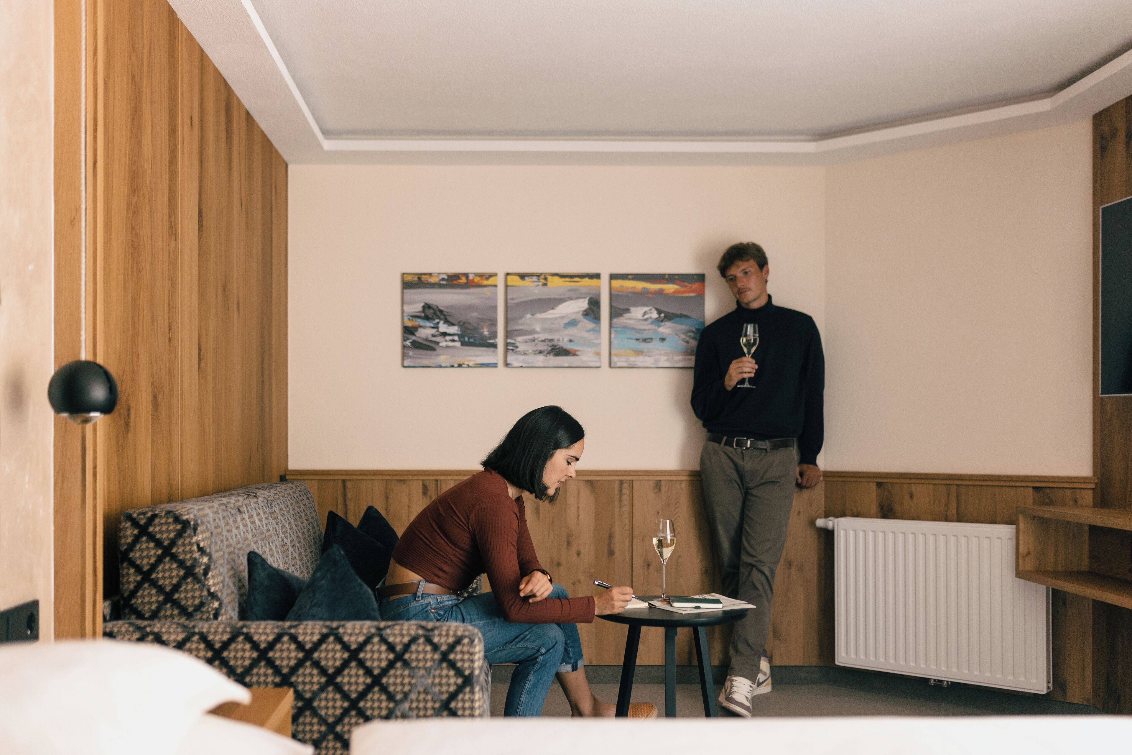 A modern hotel room with a wooden accent wall, a patterned sofa, a small round table with a glass of wine, and a wall-mounted TV.