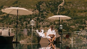 Un espace aménagé au bord de la piscine où deux personnes se détendent à l’ombre de parasols, près d’une chaise et d’un lampadaire.
