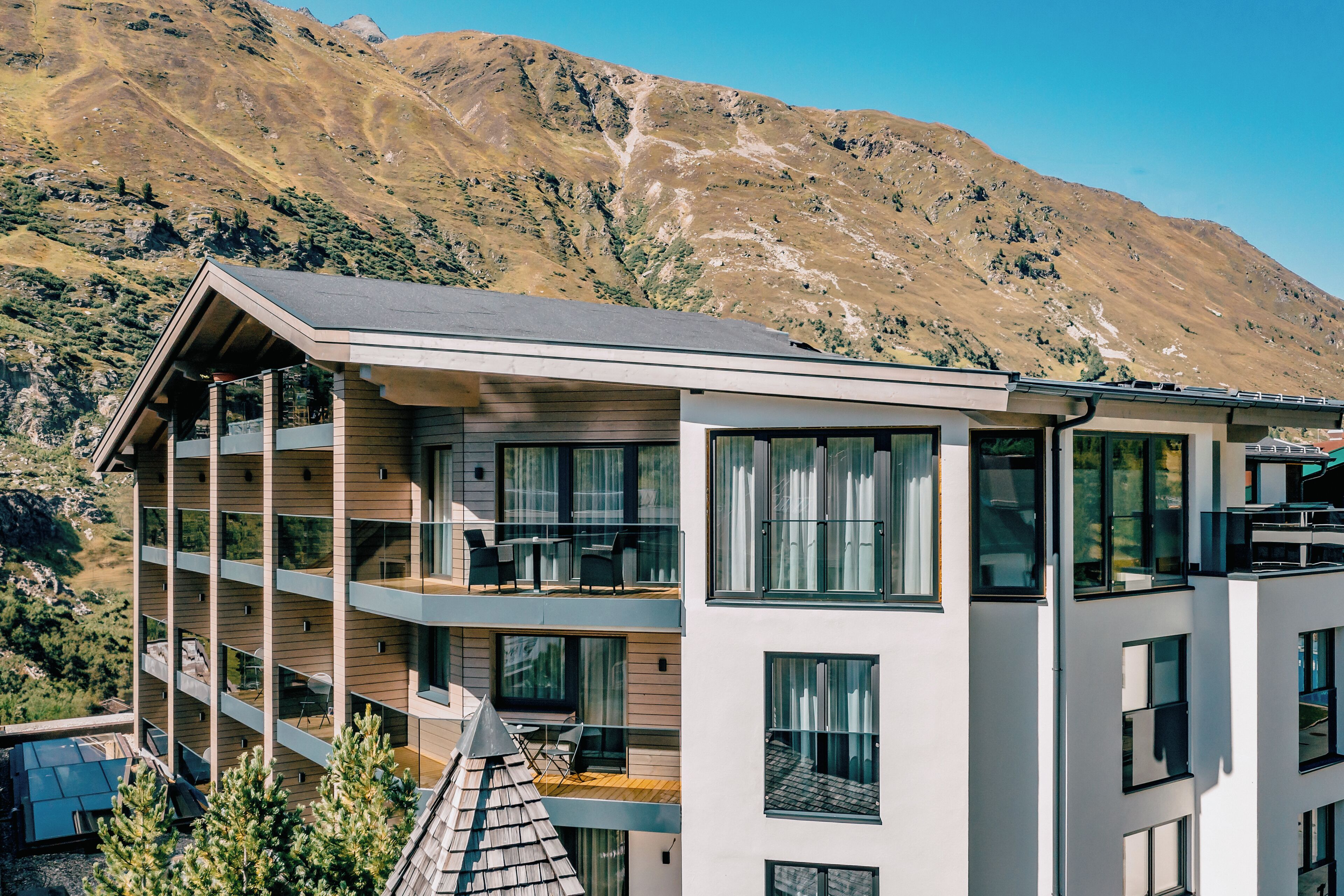 A modern multi-story building with balconies, large windows, and a mountainous backdrop.