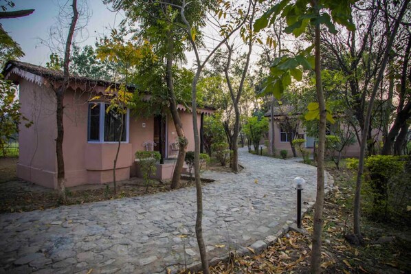 A stone-paved courtyard with a pink building, surrounded by trees and shrubs. - Ashoka Tiger Trail By VistaLux (Ramnagar)