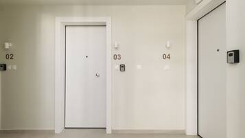 A hallway with four doors labeled 02, 03, 04, and 05. Each door has a light switch and a wall-mounted intercom. The floor is tiled, and the walls are plain.