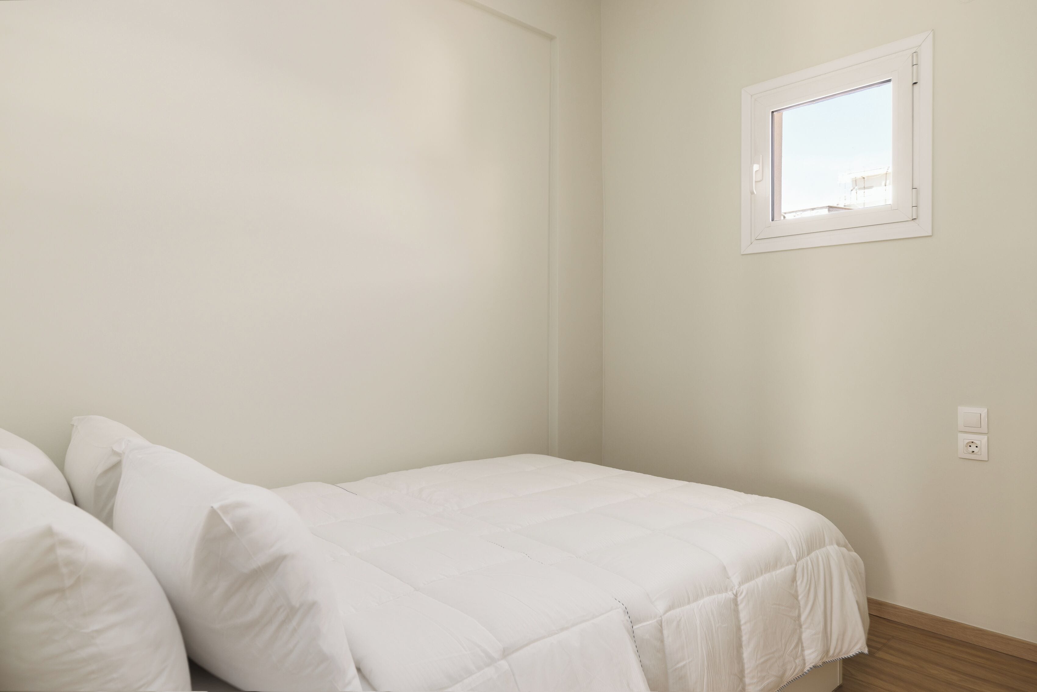 A bedroom with a single bed, white bedding, a small window, and a wall socket.