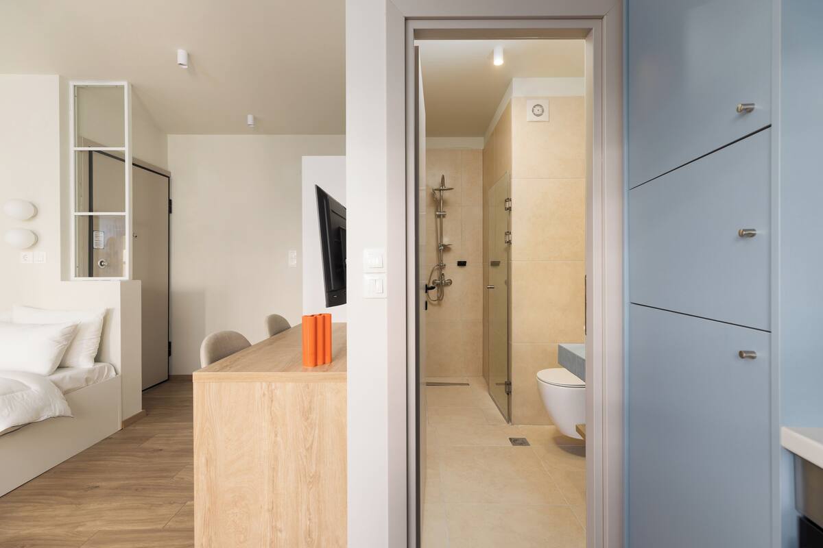 Et moderne hotelværelse med trægulv, en hvid seng, et lille træskrivebord og et bruseområde med glasdør.