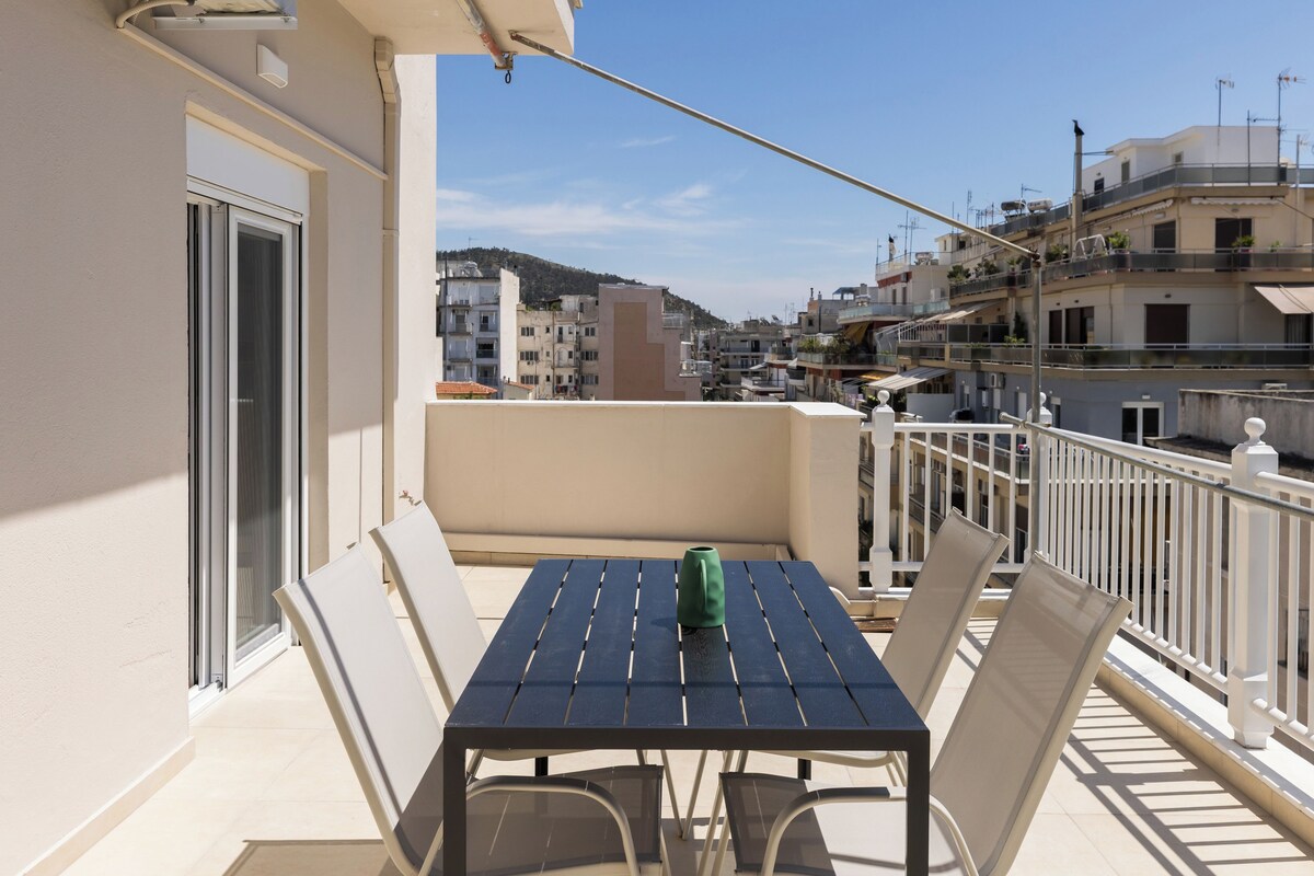 Ein Balkon mit Tisch und Stühlen, eine grüne Flasche und Blick auf eine Stadtlandschaft mit Gebäuden und einem Hügel.
