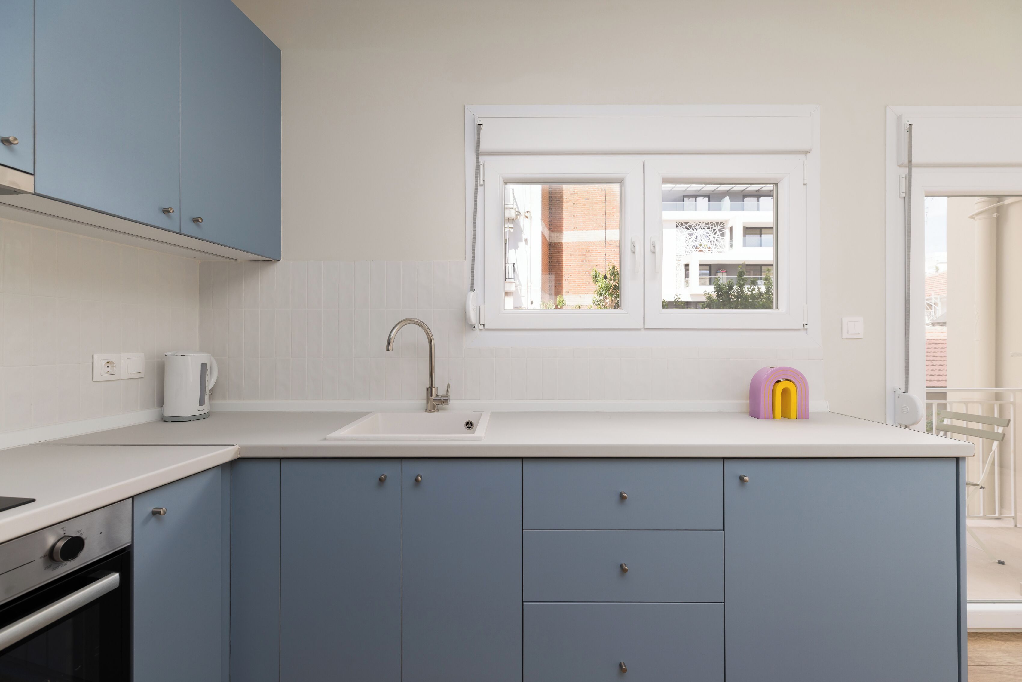 A modern kitchen with a white countertop, blue cabinets, a window, and a kettle.