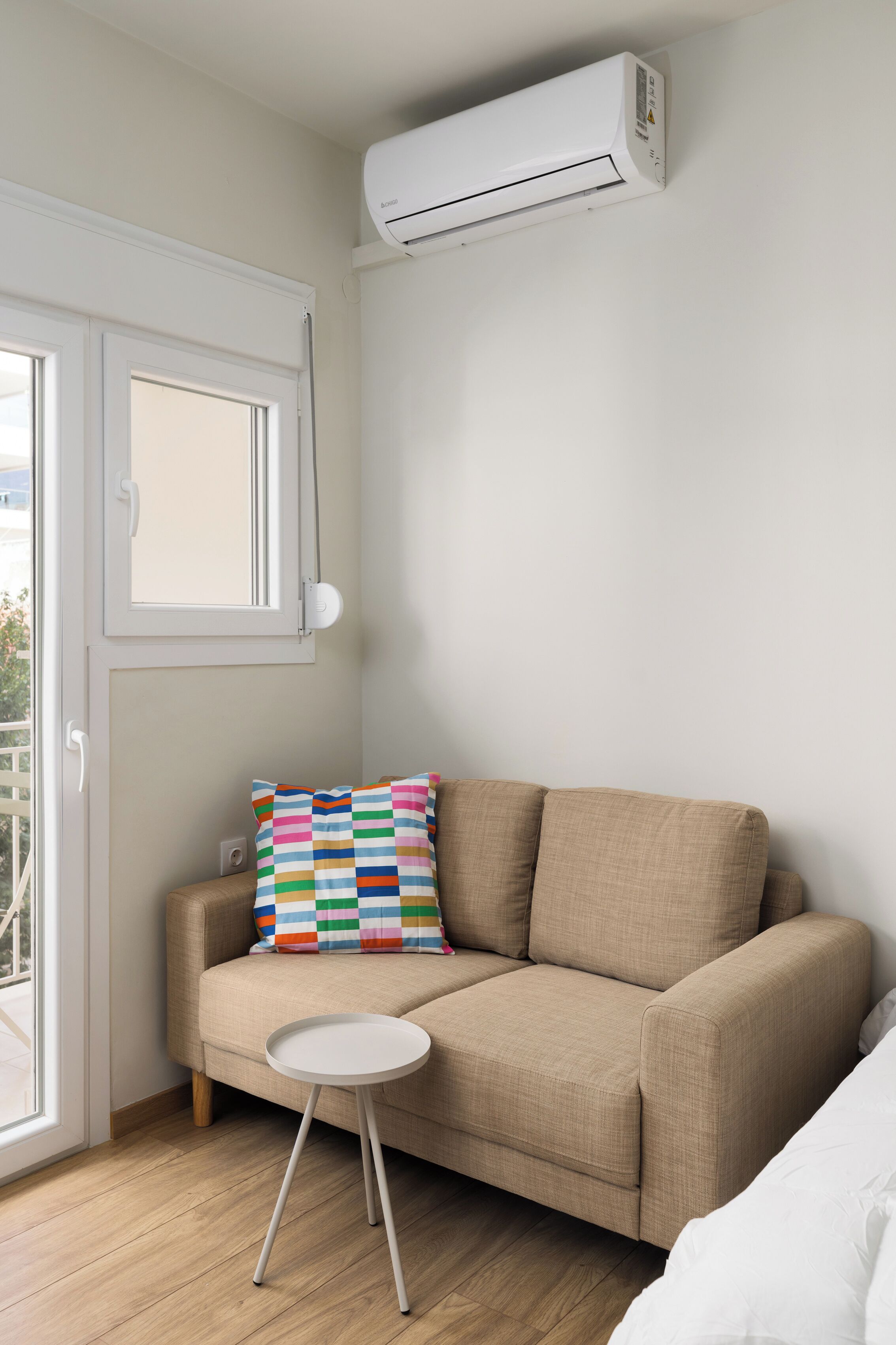 A small, modern room with a beige sofa, a colorful striped pillow, a small white side table, and an air conditioning unit mounted on the wall.