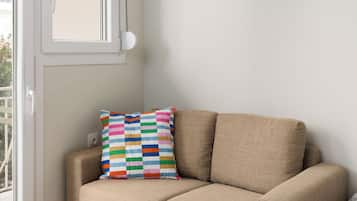 A small, modern room with a beige sofa, a colorful striped pillow, a small white side table, and an air conditioning unit mounted on the wall.