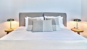 A neatly made bed with a grey headboard, white bedding, and grey and white pillows. There are two bedside tables with lamps on either side of the bed.