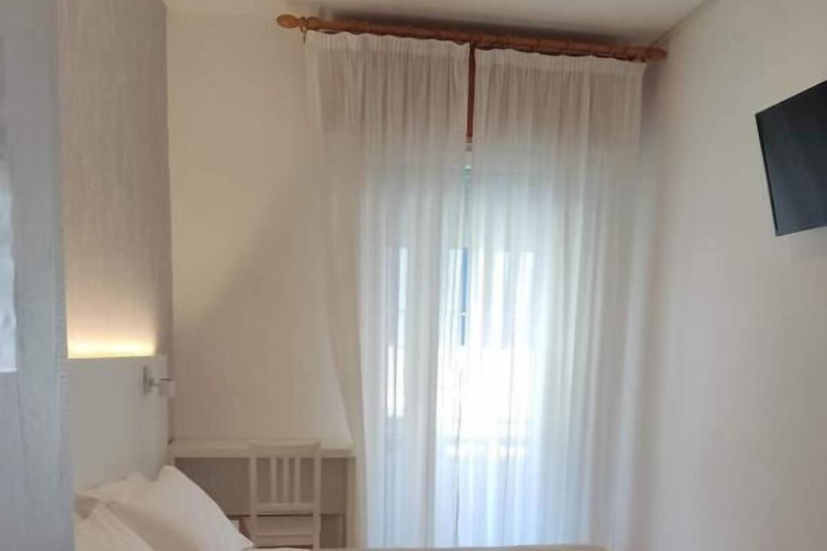A bedroom with a bed, a nightstand, a TV mounted on the wall, and a window with curtains.