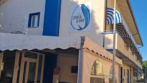 A building with a blue and white facade, a sign reading "CONCA D'ORO", outdoor seating with wooden chairs and tables, and a covered patio area. - Hotel Conca d'Oro (Rimini)