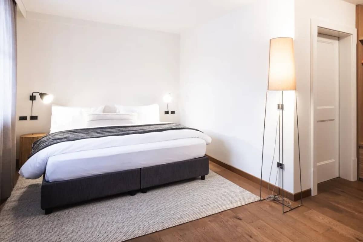 Quarto de hotel moderno com cama, abajures, um candeeiro de chão, guarda-roupa de madeira e uma porta branca.