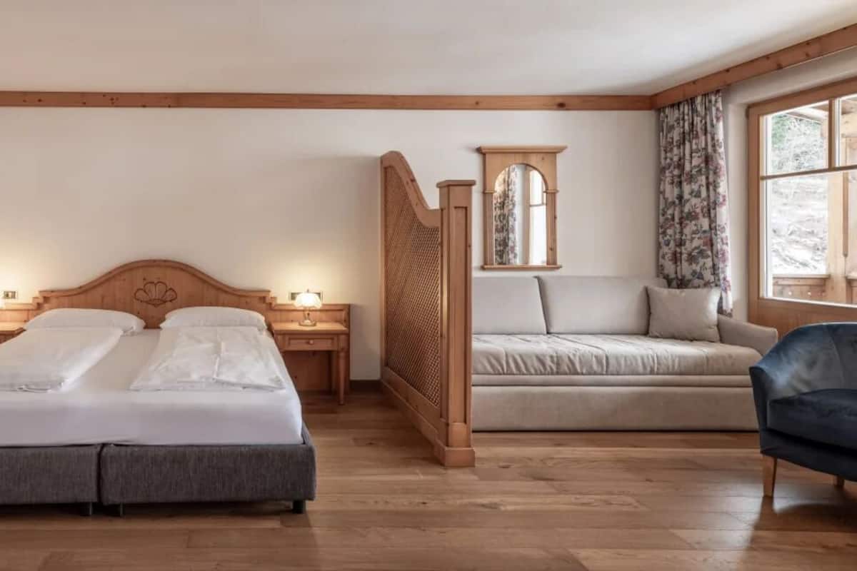 Quarto com duas camas, um sofá, uma cadeira, um separador de madeira e uma janela com cortinas.