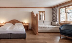 A room with two beds, a sofa, a chair, a wooden partition, and a window with curtains. - Hotel Bad Waldbrunn (Monguelfo)