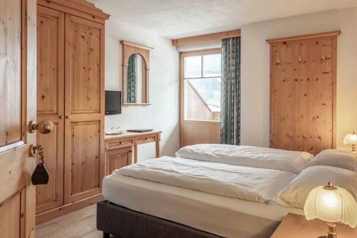 Quarto com cama de casal, guarda-roupa de madeira, escrivaninha com cadeira, espelho e janela com cortinas.