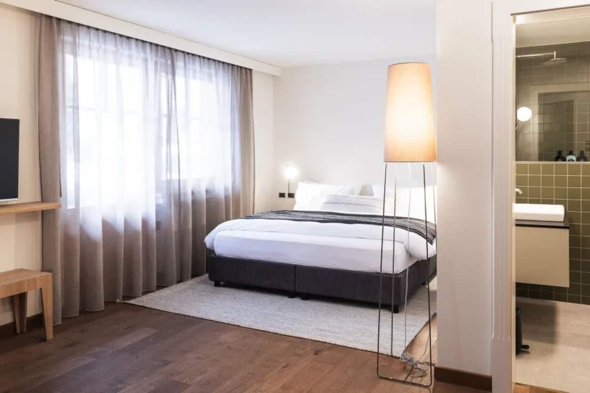 Quarto de hotel com uma cama, abajur, televisão, um banco de madeira e banheiro com espelho e pia.