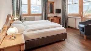 Een slaapkamer met een groot bed, houten vloer, een bureau met lamp, een stoel, een televisie en uitzicht op bergen door de ramen.