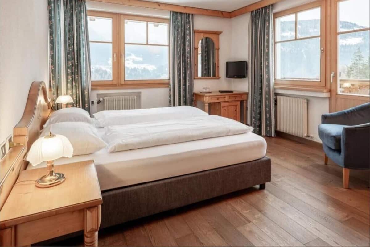 Quarto com uma cama grande, piso de madeira, uma escrivaninha com abajur, uma cadeira, uma televisão e vista para montanhas pelas janelas.