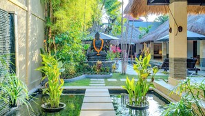 A serene garden with a Buddha statue, a thatched-roof structure, and a pool area.