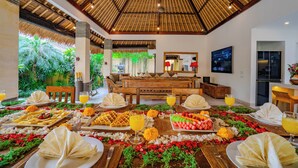 A dining area with a thatched roof, a wooden bench, and a buffet table with various dishes and drinks.