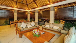An indoor lounge area with wooden furniture, a central coffee table with a floral arrangement, and a thatched roof ceiling.