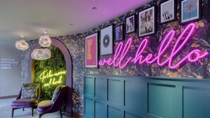Un pasillo bien iluminado con un letrero de neón que dice 'well hello', una pared verde con un cartel que dice 'For the moon and back', y una zona de estar con una silla morada.
