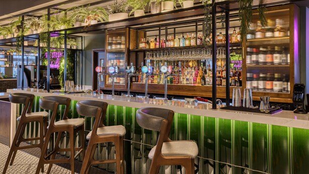 Zona de bar con taburetes, barra y estanterías llenas de botellas y vasos.