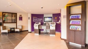 Un espace d’accueil d’hôtel avec un mur violet, un écran numérique, un panneau d’affichage pour les clients et un kiosque en libre-service.
