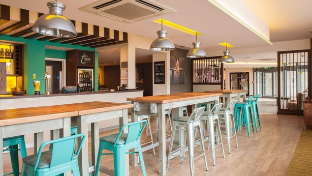 Un espace bar avec des tables en bois et des chaises de couleur turquoise, un comptoir de bar agrémenté d’un néon, et un escalier avec une rampe.