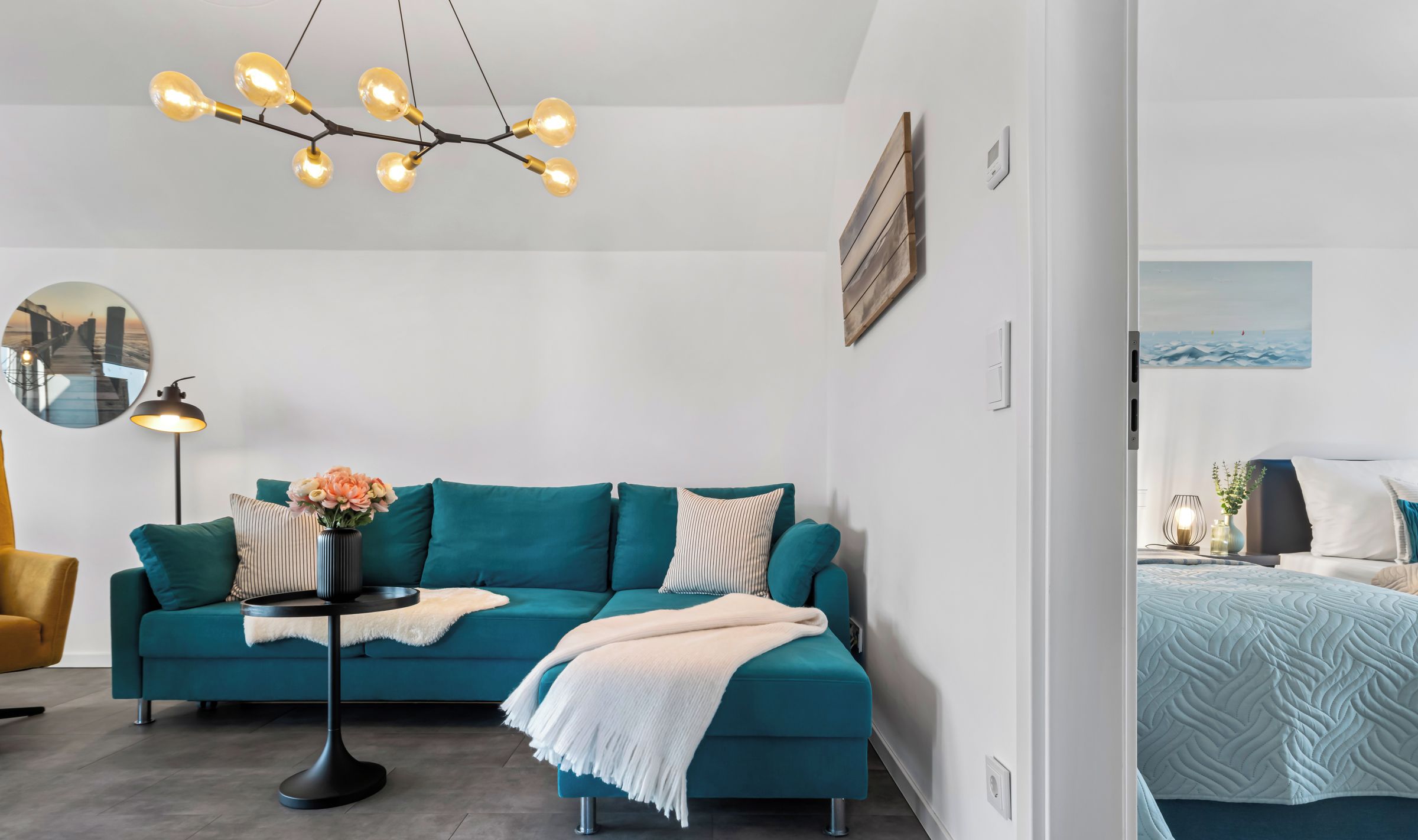 A modern living room with a teal sofa, a black side table with a vase of flowers, a hanging light fixture, and a bed with a patterned bedspread.