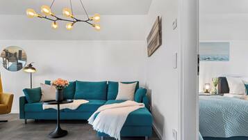 A modern living room with a teal sofa, a black side table with a vase of flowers, a hanging light fixture, and a bed with a patterned bedspread.