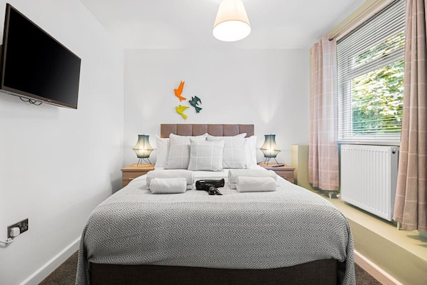 A hotel room with a bed, two bedside tables, a TV mounted on the wall, a window with curtains, and a radiator.