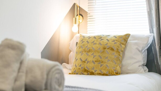 A neatly made bed with a patterned pillow, a rolled towel, and a modern lamp.