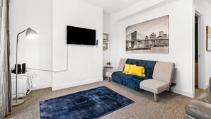 A modern living room with a flat-screen TV, a sofa with a throw blanket, a blue rug, and a cityscape painting on the wall.