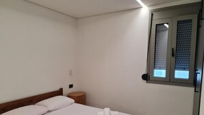 Een slaapkamer met een bed, nachtkastje, houten vloer en een raam met jaloezieën.