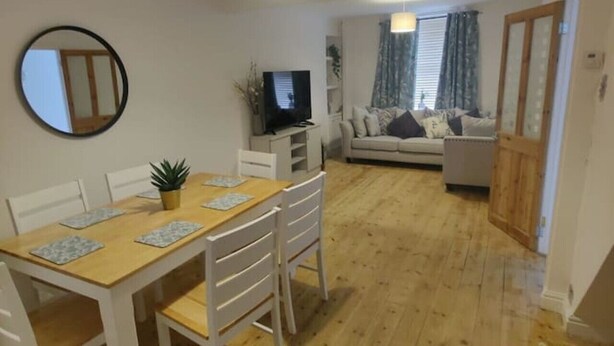 A dining area with a wooden table and chairs, a television, and a sofa in the background. - 3 Bed • Parking • Smart TV`s • Great for Work (Clydach)