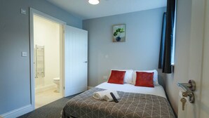 A bedroom with a bed, a door, a towel, a remote, and a painting on the wall. - Contractor Stay • 2 Bed • 2 En-suites (Swansea)